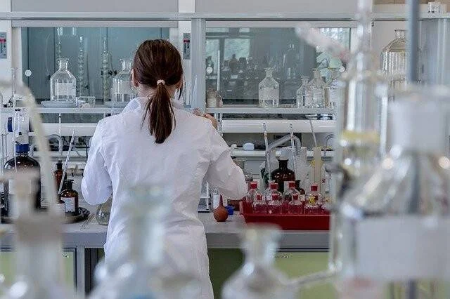 Mujer con bata blanca de espaldas trabaja en una mesada de laboratorio rodeada de frascos y reactivos.