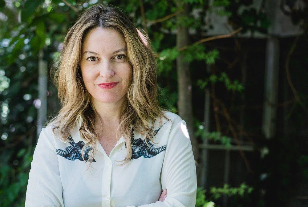 Mujer de cabello rubio ondulado, con blusa blanca y brazos cruzados, posando sonriente ante un fondo de vegetación.