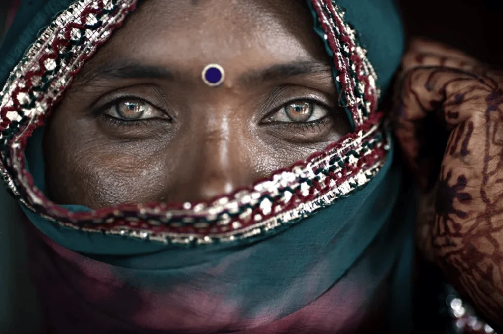 Primer plano del rostro de una mujer con velo de sari verde y bindi azul, mostrando llamativos ojos de color claro.