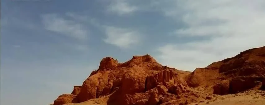 Acantilados de roca rojiza erosionada se elevan sobre un paisaje desértico bajo un cielo azul con nubes dispersas.
