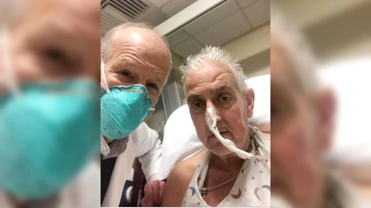 Un médico con mascarilla quirúrgica posa junto a un paciente adulto mayor con cánula nasal en una habitación de hospital.