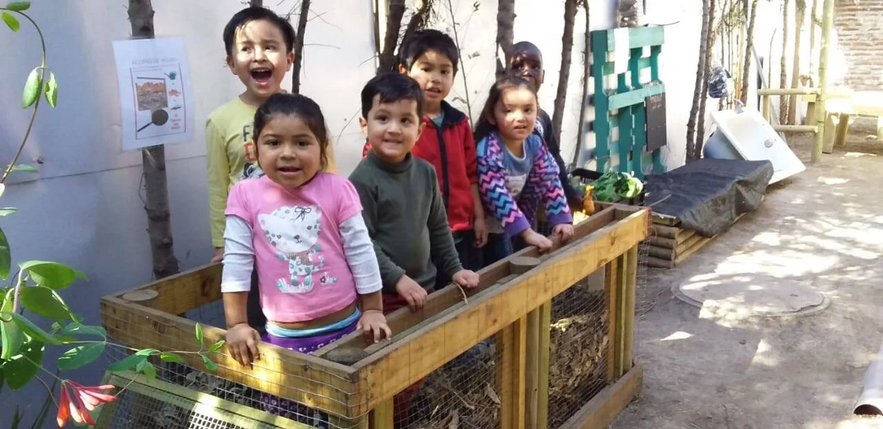 Grupo de niños en edad preescolar posan sonrientes junto a una huerta elevada de madera en un patio escolar.