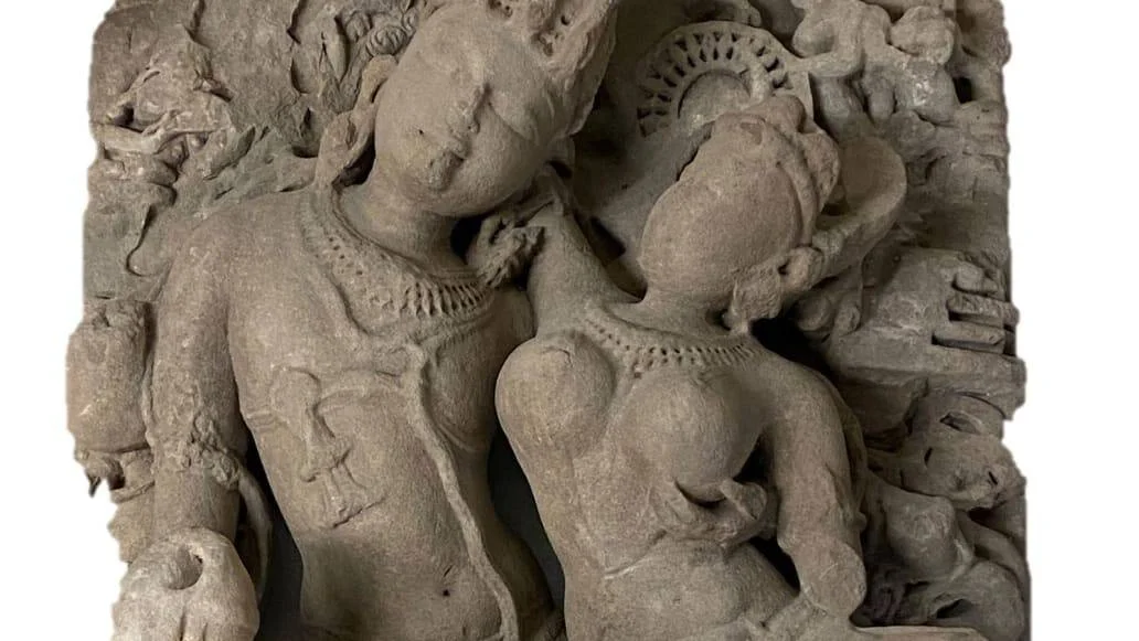 Relieve en piedra gris con dos figuras humanas adornadas con collares y tocados, estilo escultórico del arte clásico indio.