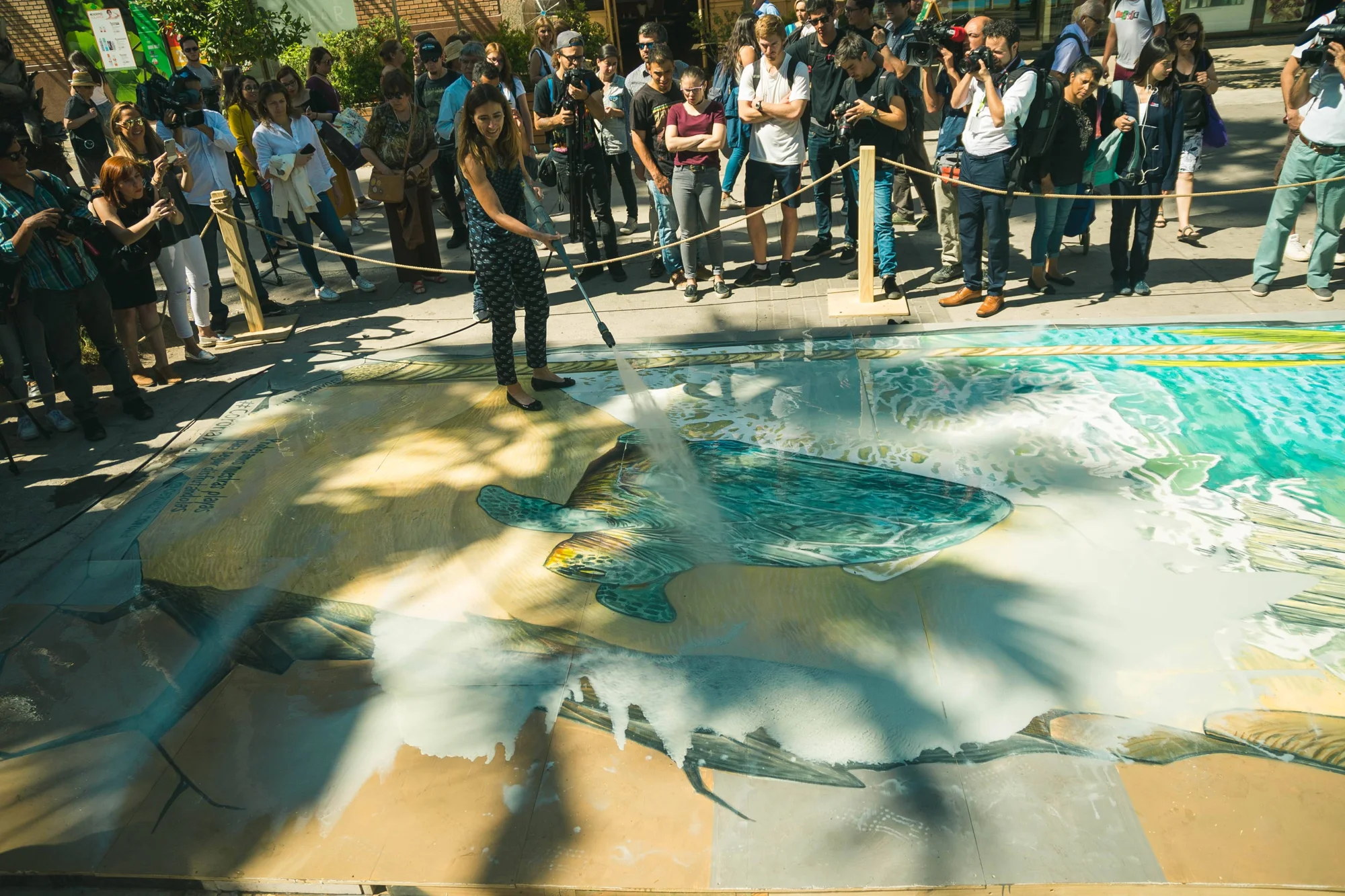 Una mujer rocía agua sobre un gran mural 3D de peces marinos pintado en el suelo, rodeada de público y fotógrafos.
