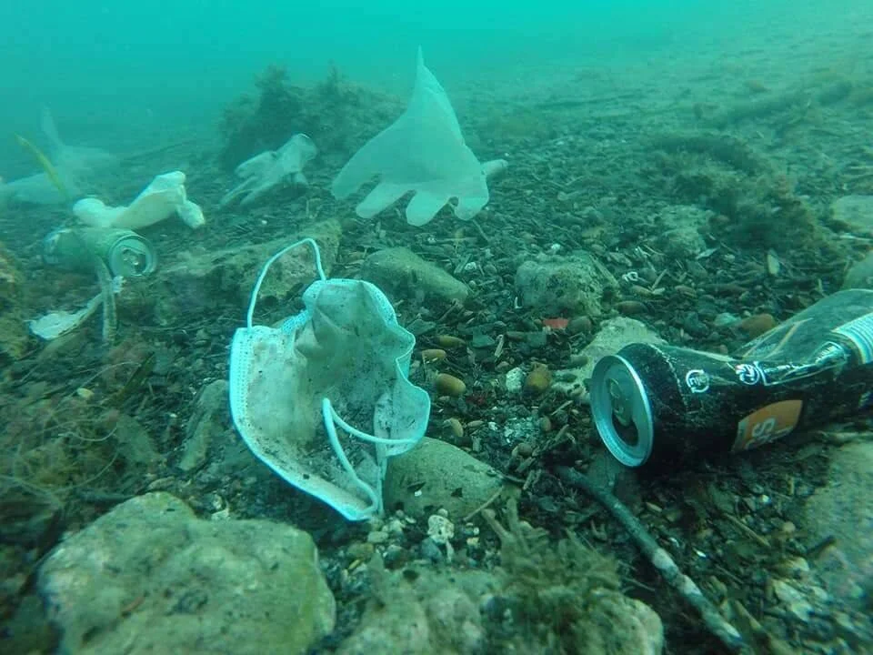 Mascarilla quirúrgica, latas, bolsas plásticas y botellas acumuladas en el fondo del mar, mostrando contaminación oceánica.