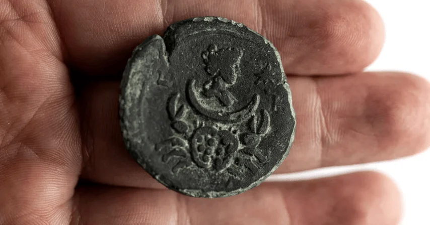 Mano sosteniendo una moneda antigua de bronce oscurecido con relieve de figura humana y motivos vegetales.