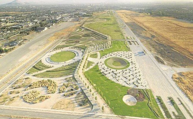 Vista aérea de un amplio parque urbano con jardines geométricos circulares y áreas verdes, rodeado de pistas y una ciudad al fondo.