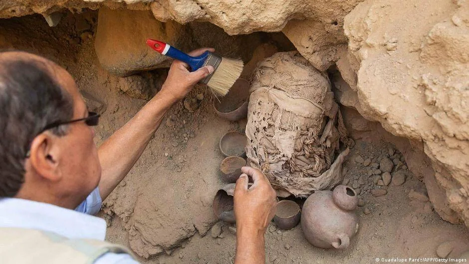 Arqueólogo cepilla con brocha una momia envuelta en telas junto a vasijas de cerámica en una tumba excavada.