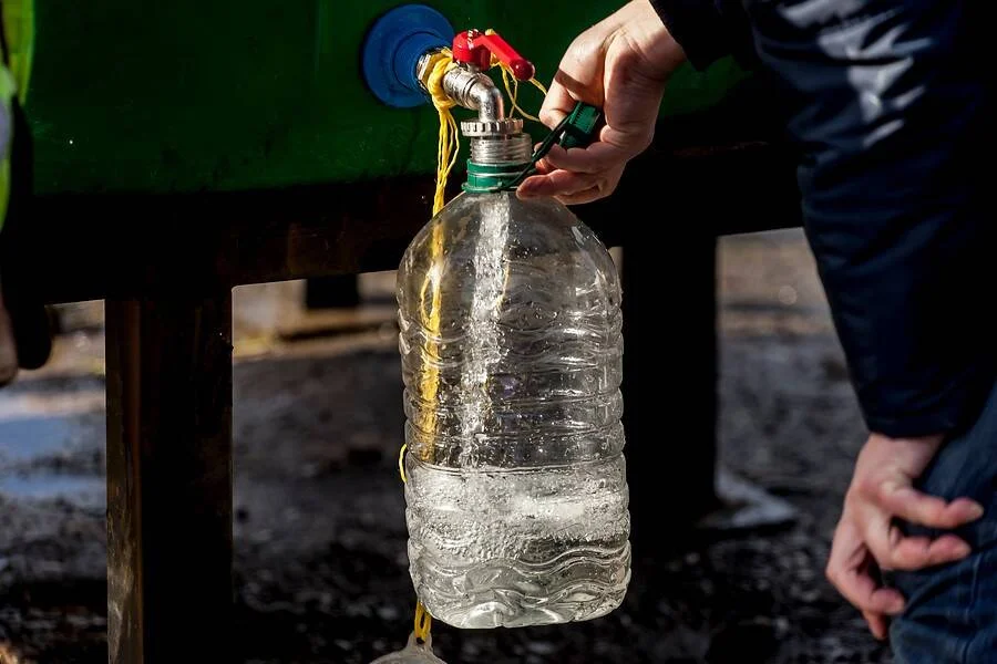 Una mano llena una botella plástica grande con agua desde un grifo conectado a un tanque verde.