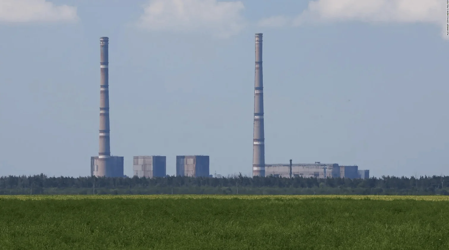 Dos altas chimeneas de una planta industrial se elevan sobre campos verdes y arboleda bajo un cielo parcialmente nublado.