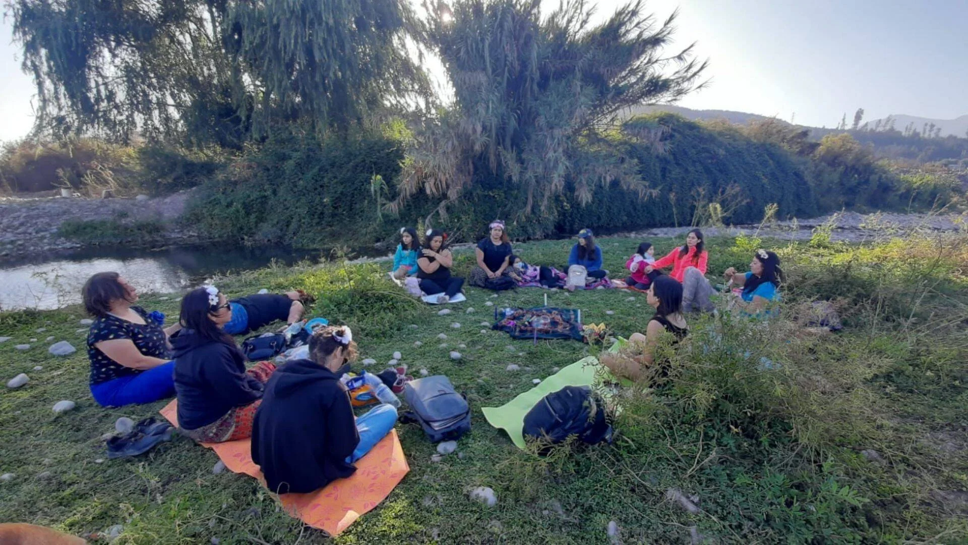Grupo de mujeres sentadas en círculo sobre el pasto a orillas de un río, participando de un encuentro al aire libre.