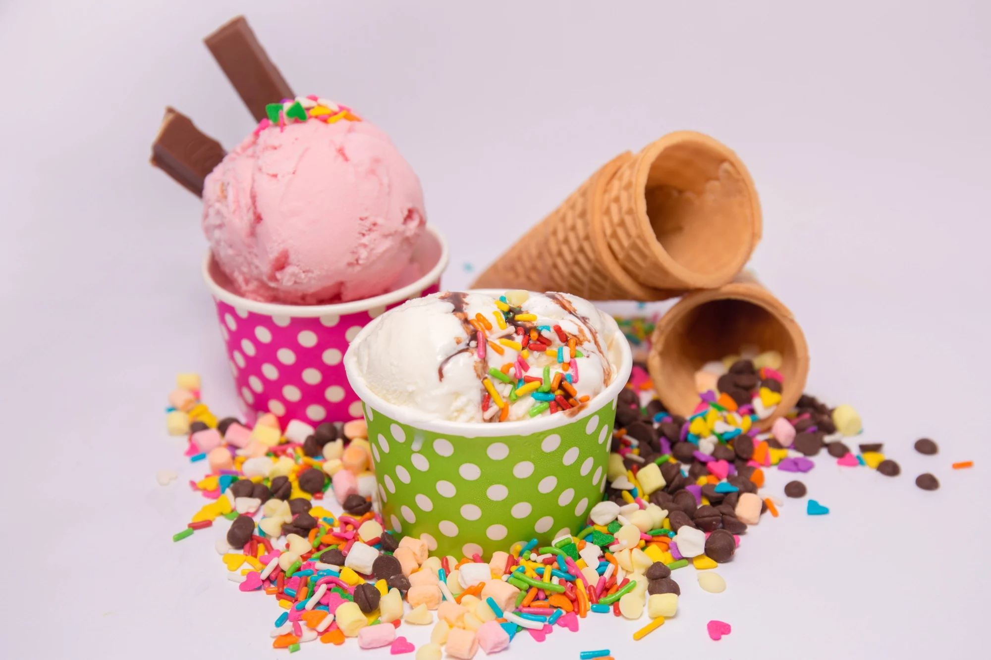 Dos vasitos de helado —uno rosa con barras de chocolate y otro blanco con sprinkles y salsa— rodeados de conos de waffle, chispas de chocolate y mini malvaviscos.