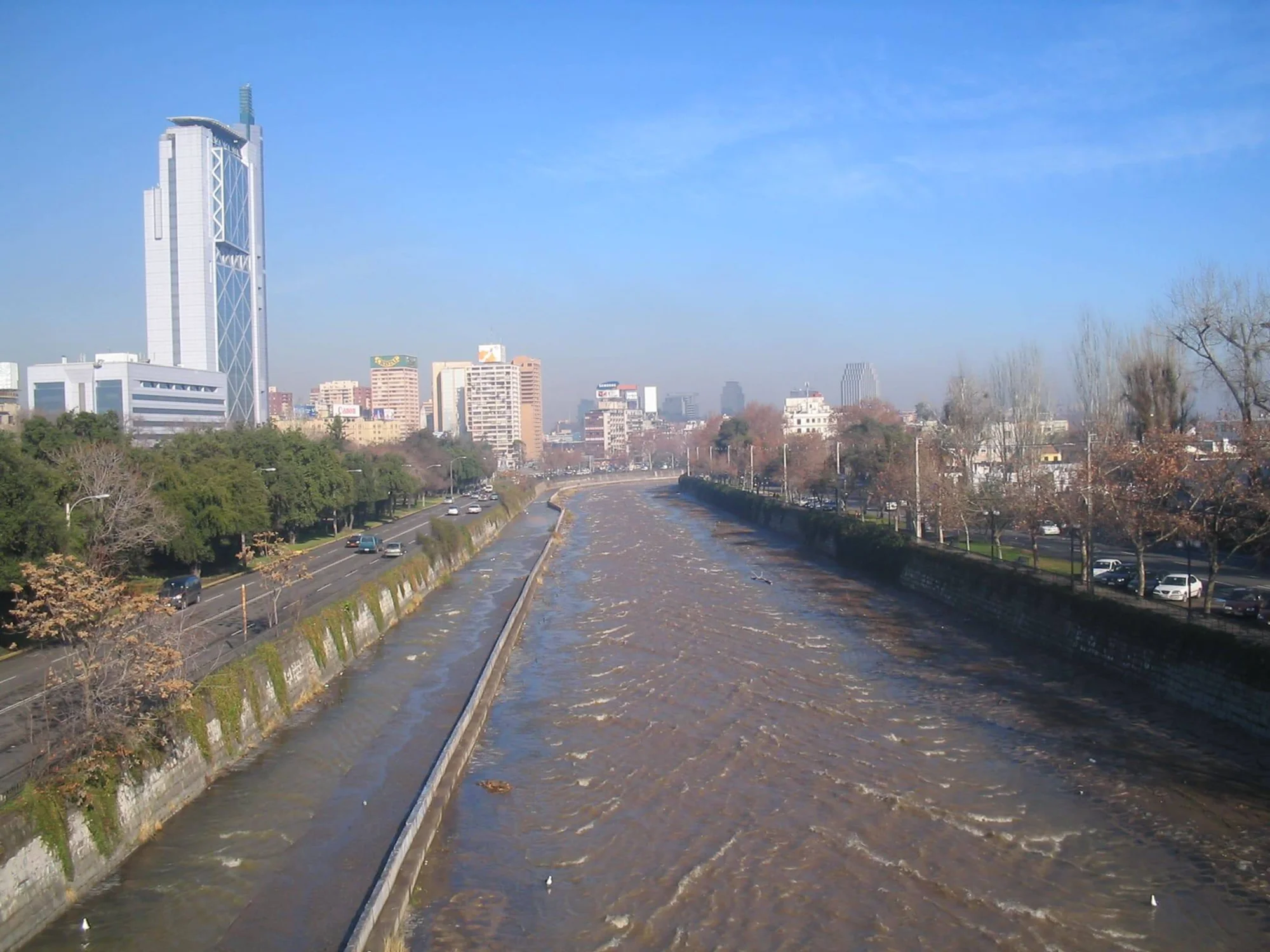 Río de aguas turbias y caudalosas atraviesa una ciudad, flanqueado por avenidas con tráfico y edificios altos al fondo.