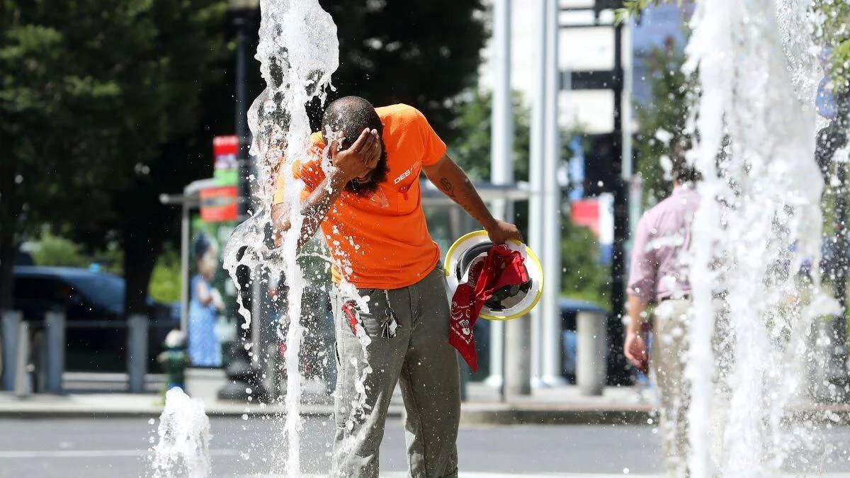 Un trabajador con camiseta naranja y casco en mano se moja la cabeza en una fuente pública para combatir el calor extremo.