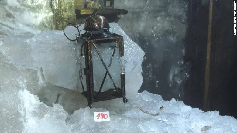 Linterna metálica oxidada rodeada de bloques de hielo, con una etiqueta numerada, en exhibición del Museo de la Guerra Blanca.
