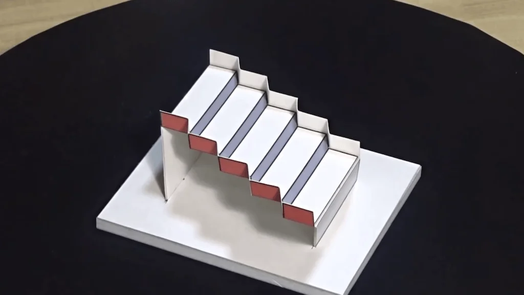 Maqueta arquitectónica de una escalera construida con papel blanco, gris y rojo, sobre una base blanca en mesa oscura.