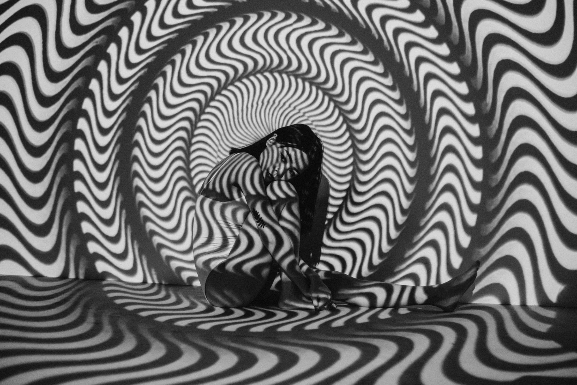 Persona sentada en el suelo cubierta por proyecciones de líneas onduladas en blanco y negro que crean un efecto óptico hipnótico.