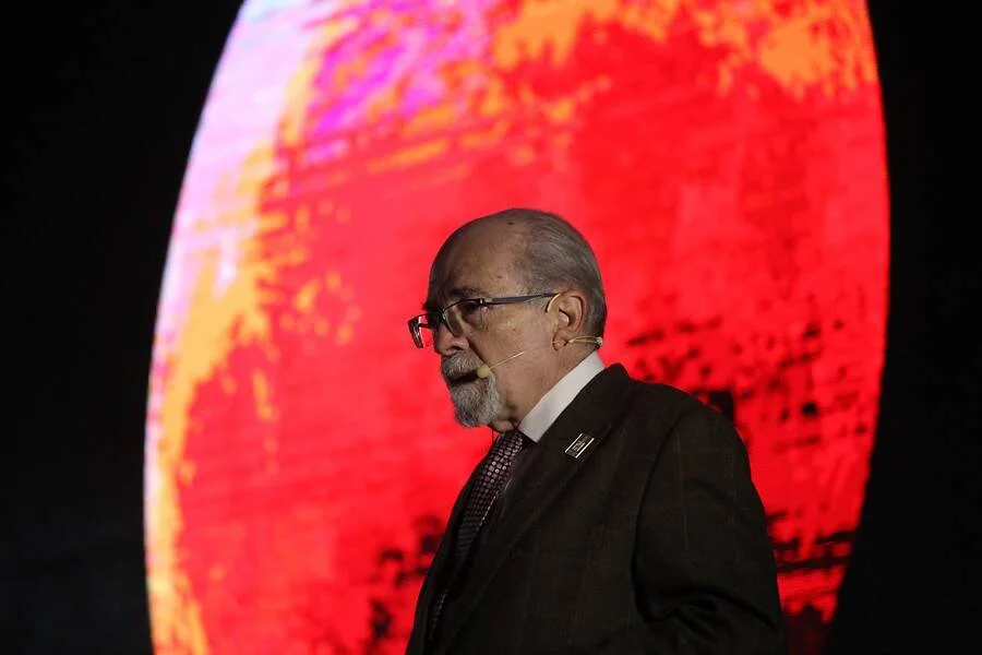 Hombre mayor con traje oscuro y micrófono de solapa habla frente a una gran pantalla con imagen en tonos rojos y naranjas.