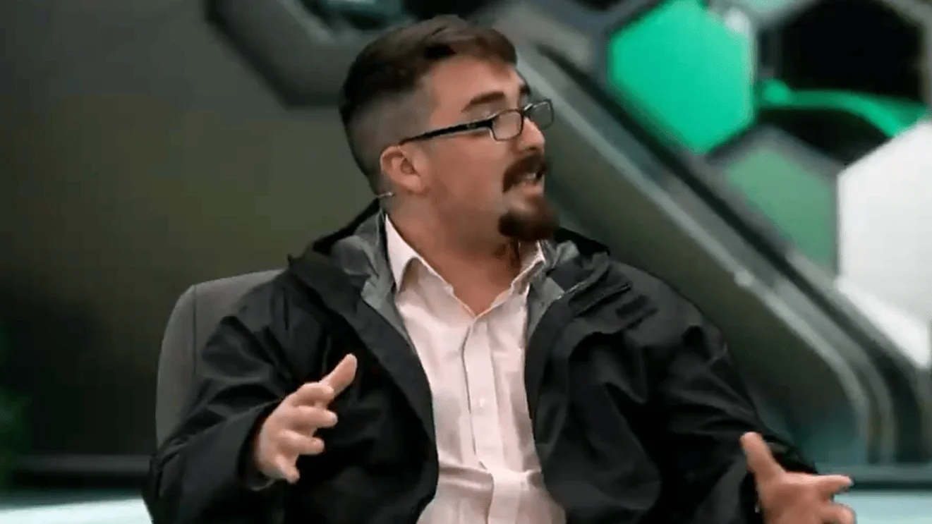 Hombre joven con gafas y barba gesticula con las manos mientras habla en un estudio de televisión.