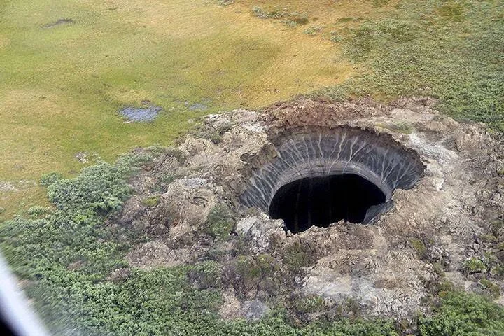 Vista aérea de un enorme cráter circular en la tundra, con paredes verticales de tierra y roca que descienden hacia una cavidad oscura.