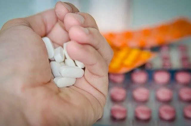 Una mano abierta sostiene varias pastillas blancas, con blísteres de medicamentos rosados y naranjas desenfocados al fondo.