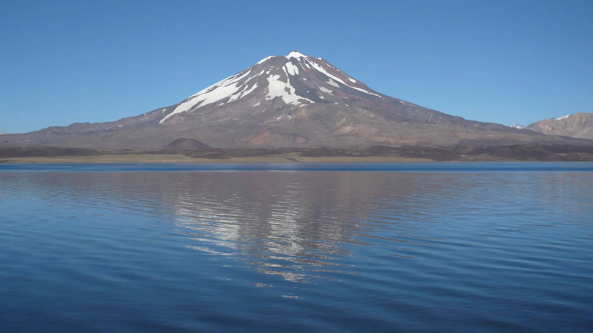 Volcán de cima nevada se refleja sobre las aguas tranquilas de un lago andino bajo cielo despejado.