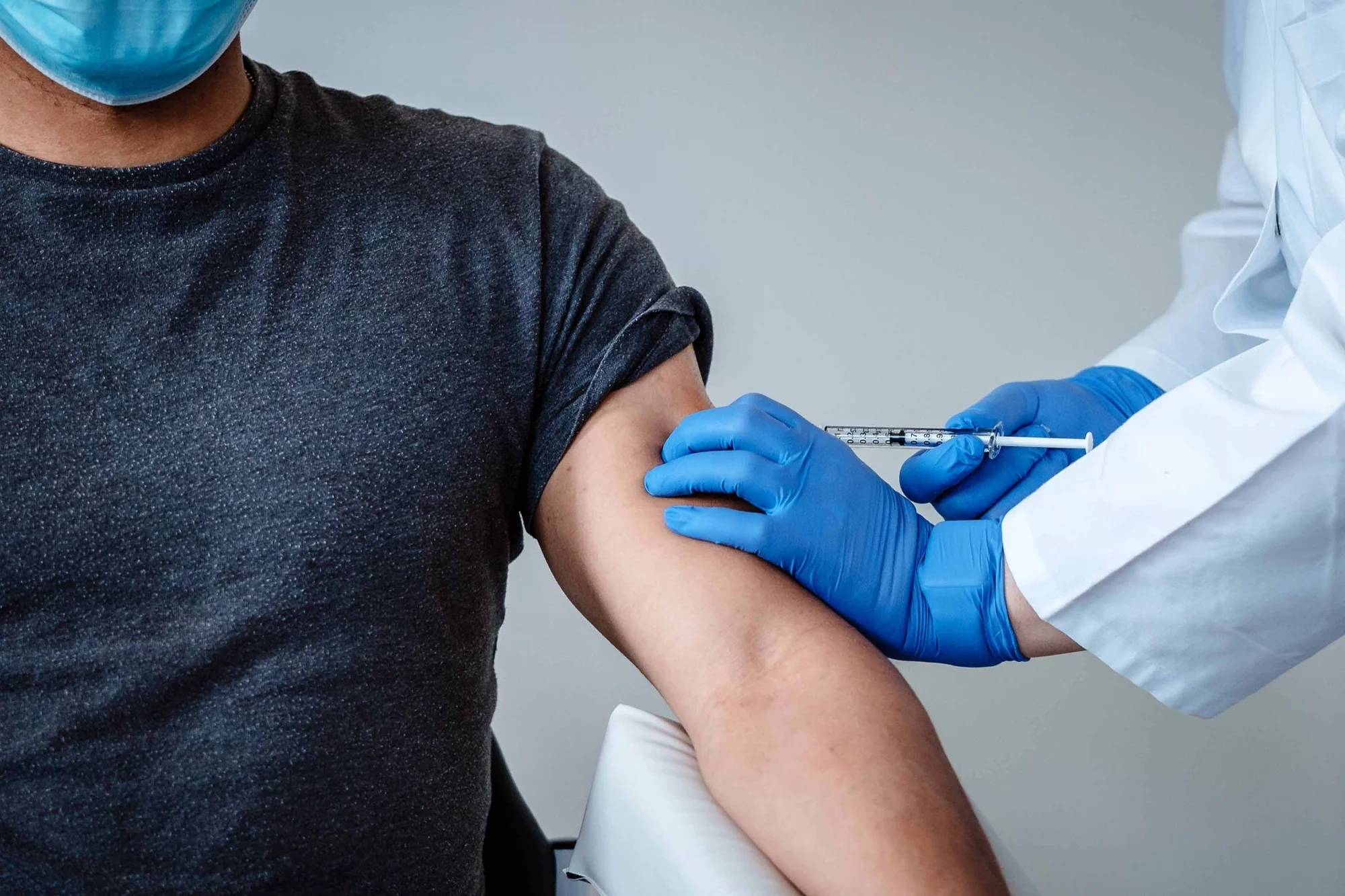 Profesional de salud con guantes azules aplica una inyección en el brazo de una persona con mascarilla quirúrgica.