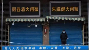 Un guardia con mascarilla custodia la entrada cerrada de un mercado en China, con letreros en chino sobre los locales.