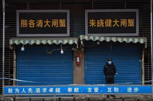 Un guardia con mascarilla custodia la entrada cerrada de un mercado en China, con letreros en chino sobre los locales.