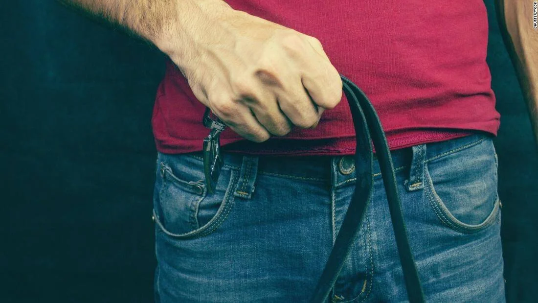 Una mano sujeta una pistola oculta bajo la camiseta, siendo extraída del cinturón de un pantalón vaquero.