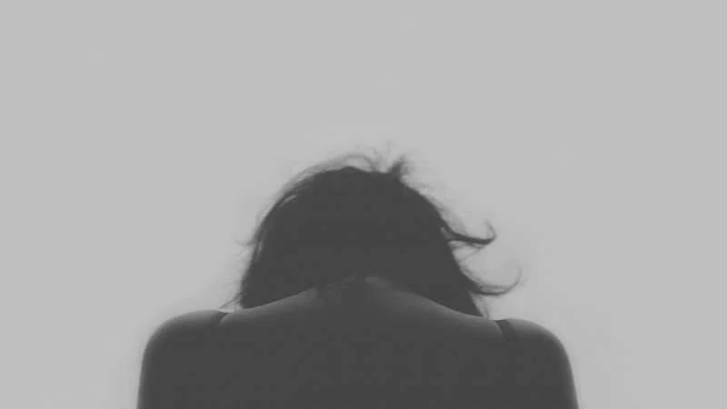 Persona vista de espaldas con la cabeza inclinada hacia abajo y el cabello al viento, en tono gris evocando tristeza o angustia.