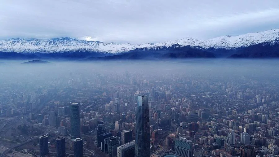 Vista aérea de una gran ciudad envuelta en una densa capa de smog, con una cordillera nevada al fondo bajo cielo nublado.