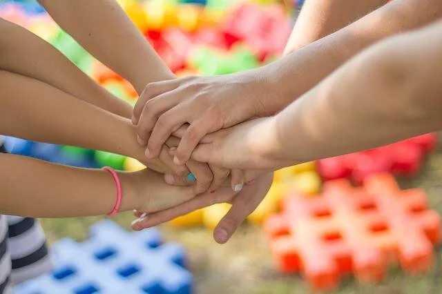 Varias manos de niños y adultos apiladas juntas en señal de unidad, con juguetes de colores de fondo.