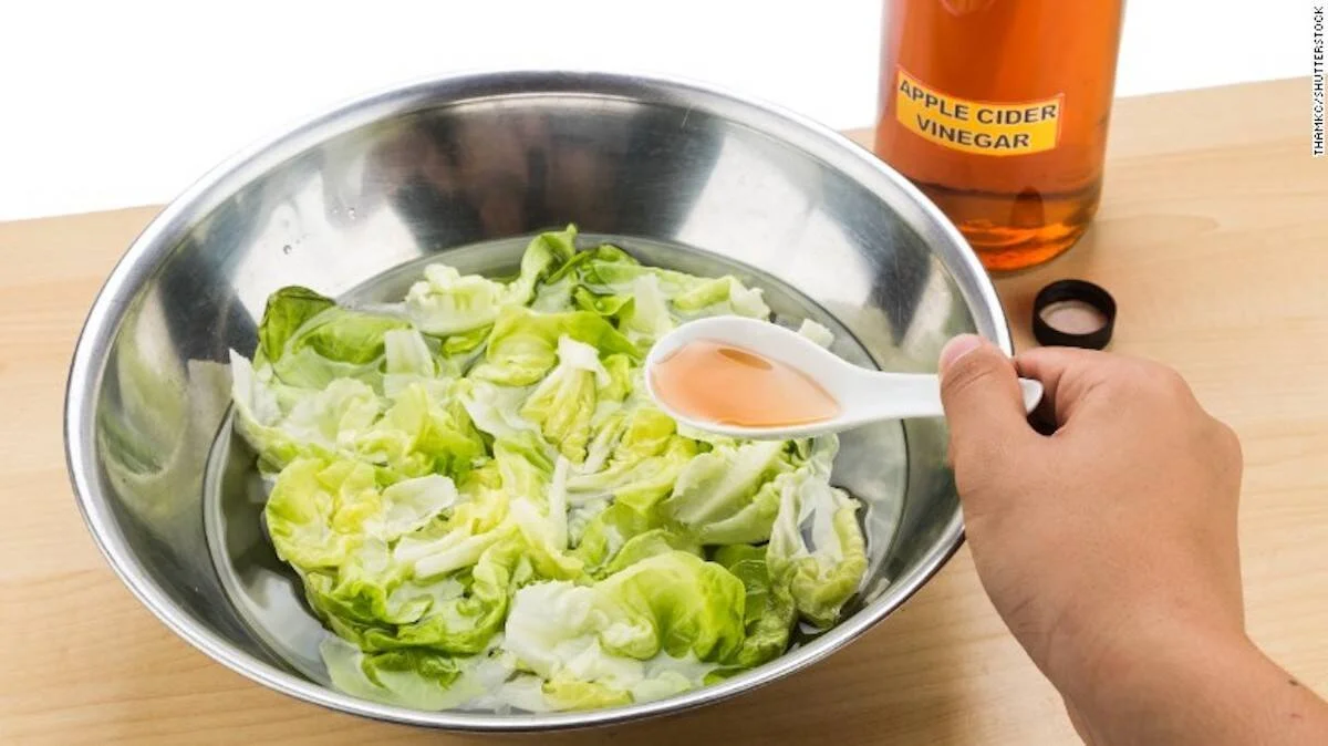 Mano vertiendo vinagre de sidra de manzana sobre ensalada de lechuga en un bol de acero inoxidable.