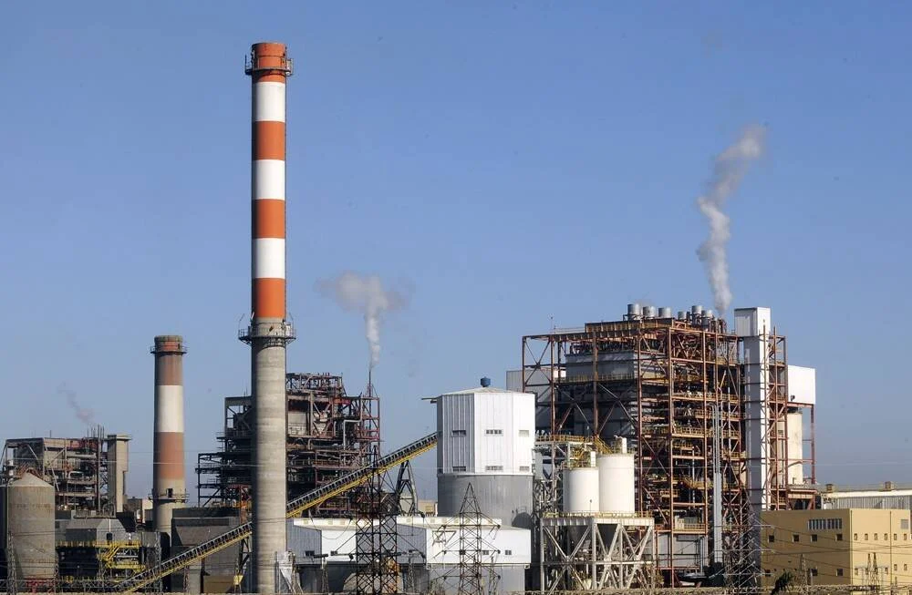 Planta industrial con chimeneas a rayas rojas y blancas expulsando humo bajo un cielo azul despejado.