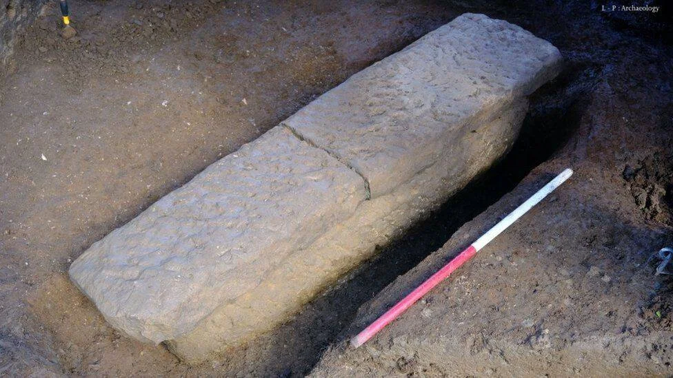 Sarcófago medieval de piedra recién excavado junto a una escala de medición roja y blanca.