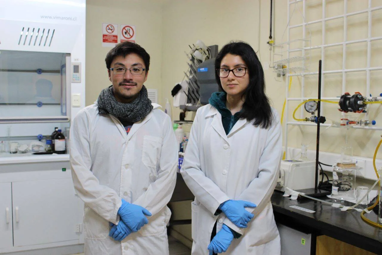 Un hombre y una mujer con batas blancas y guantes azules posan juntos en un laboratorio científico equipado.