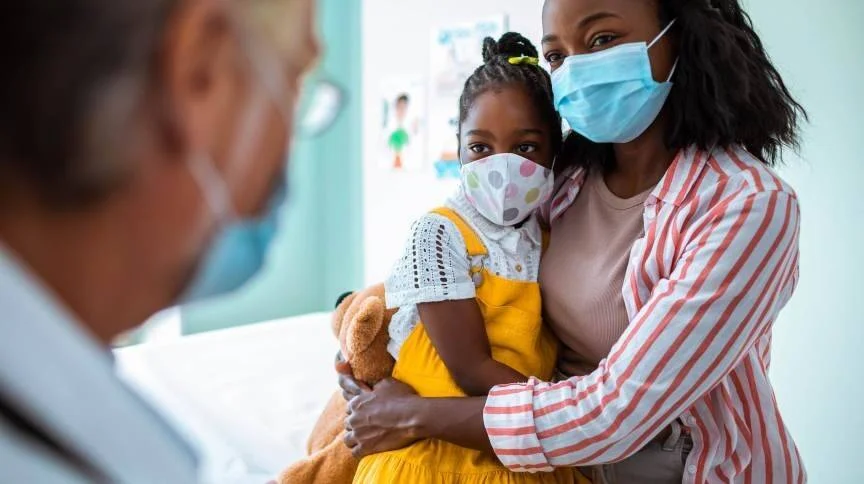 Una madre abraza a su hija pequeña, ambas con mascarilla, durante una consulta médica con un doctor.