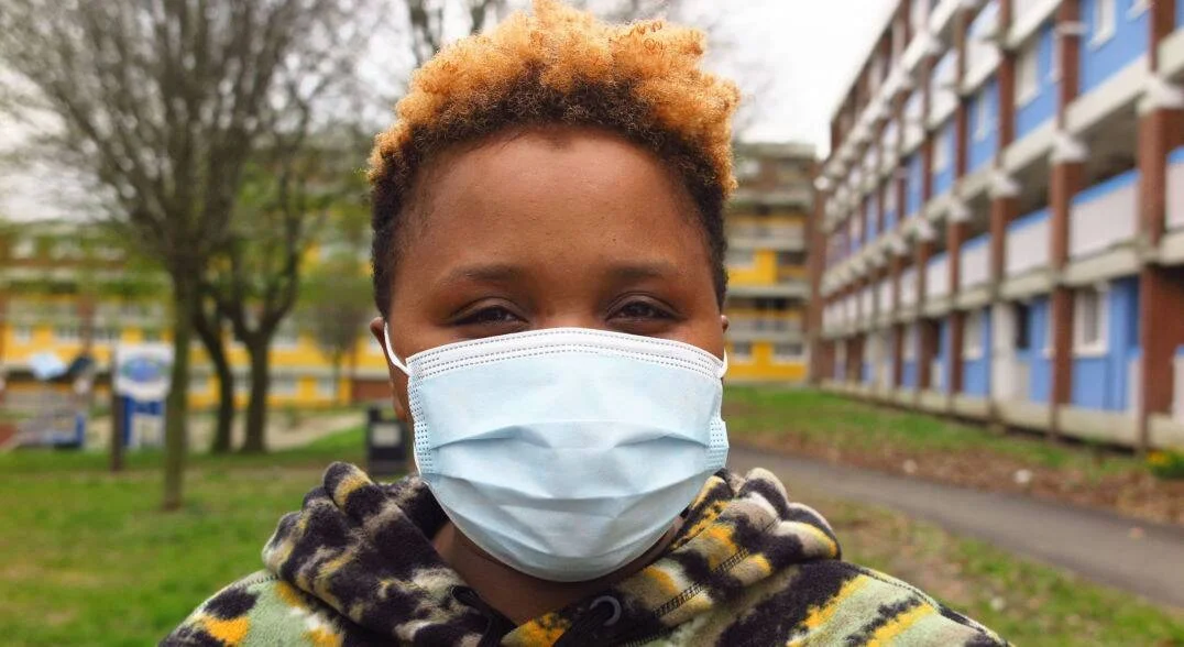 Joven con cabello teñido de rubio usa mascarilla quirúrgica azul frente a edificios residenciales de colores.