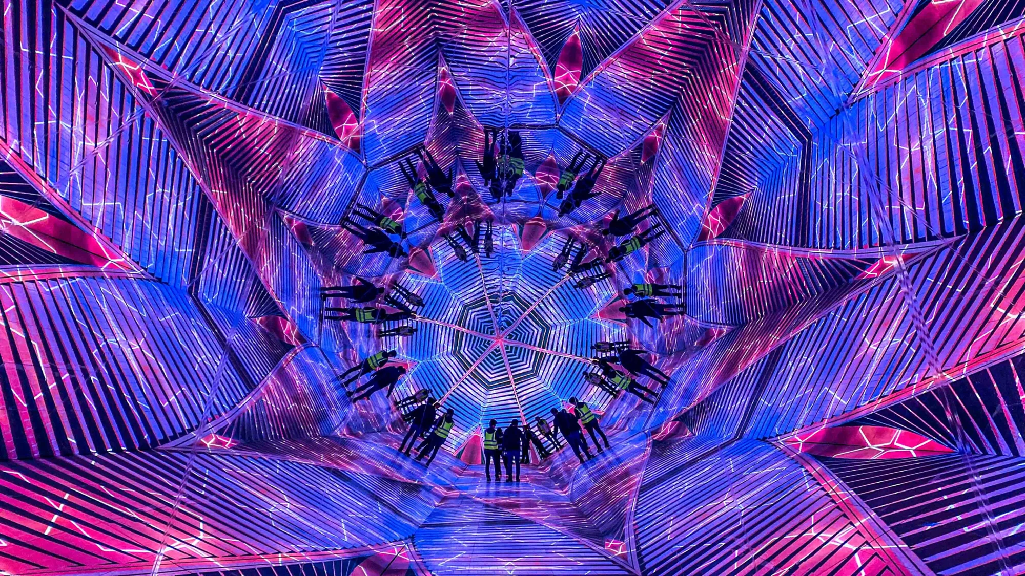 Personas con chalecos reflectantes en el interior de una instalación inmersiva con espejos y luces de neón azules y rosas que crean un efecto caleidoscópico.