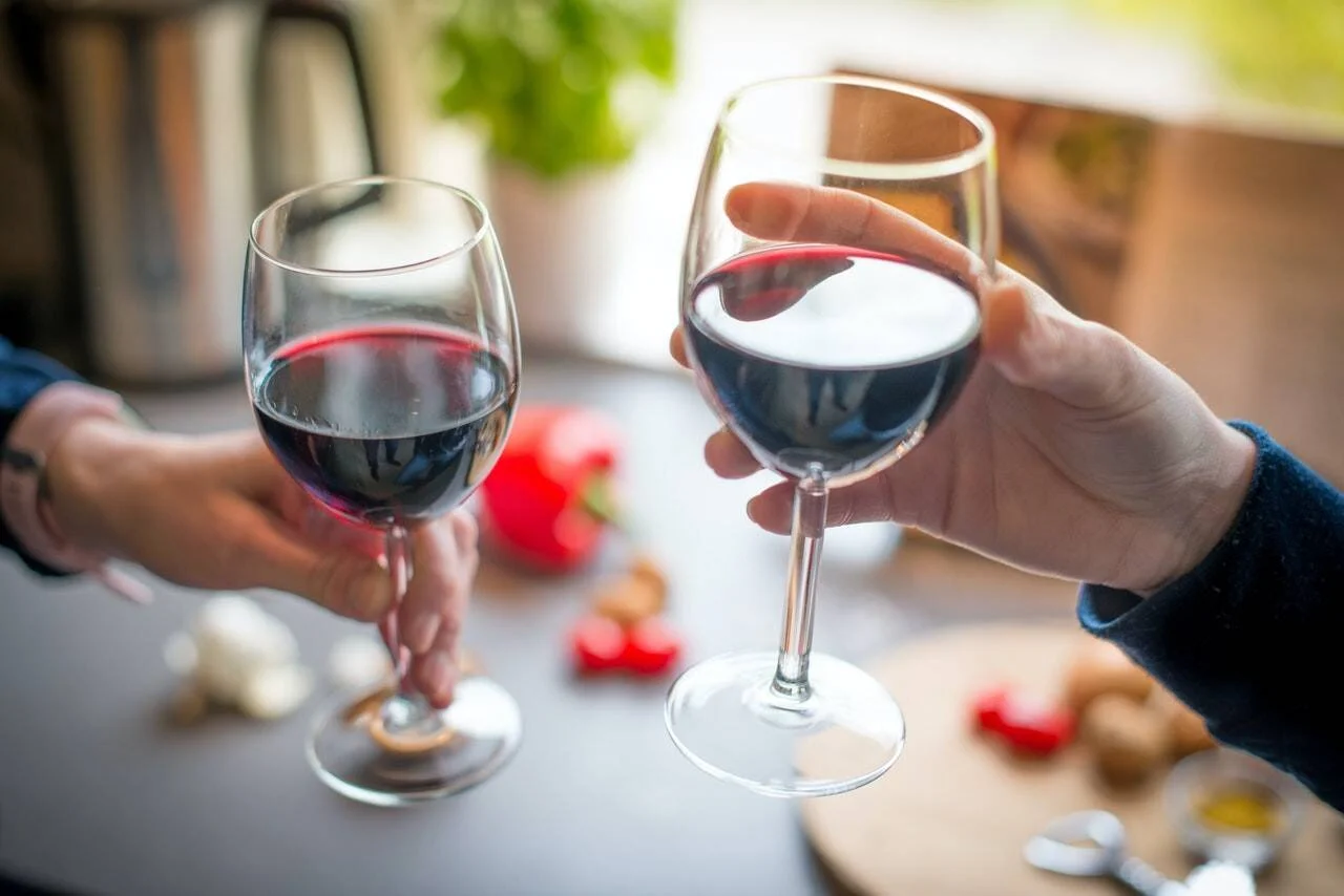 Dos manos chocan copas de vino tinto sobre una mesa con verduras y especias de fondo.