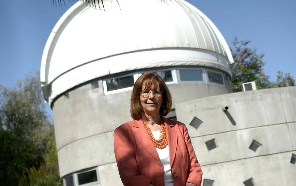Mujer con blazer rosa y collar naranja posa frente a la cúpula blanca de un observatorio astronómico en día soleado.
