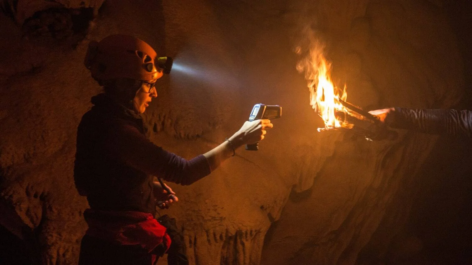 Investigadora con casco y linterna frontal apunta un termómetro infrarrojo a una hoguera encendida dentro de una cueva oscura.