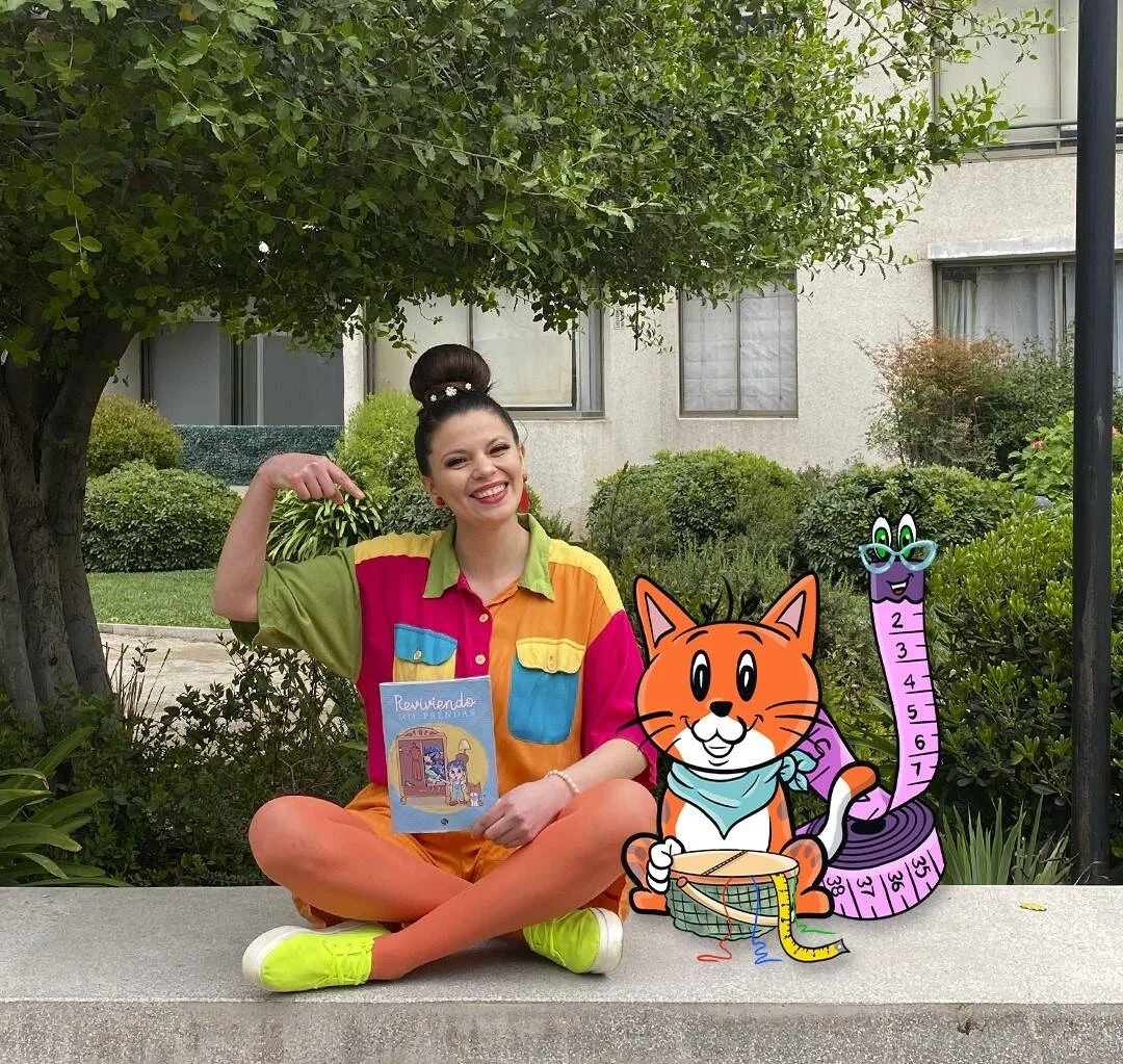 Mujer sonriente con ropa colorida sostiene el libro infantil 'Reviviendo mis prendas', junto a ilustraciones de un gato y una cinta métrica.
