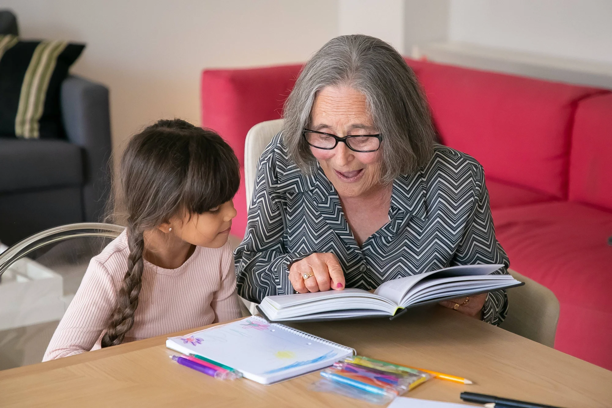 Mujer mayor con gafas lee un libro junto a una niña en una mesa con cuadernos y lápices de colores.