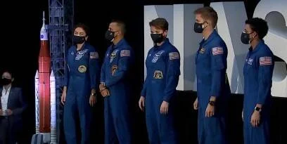 Cinco astronautas de la NASA con trajes azules y mascarillas posando junto a una maqueta del cohete SLS durante un evento oficial.