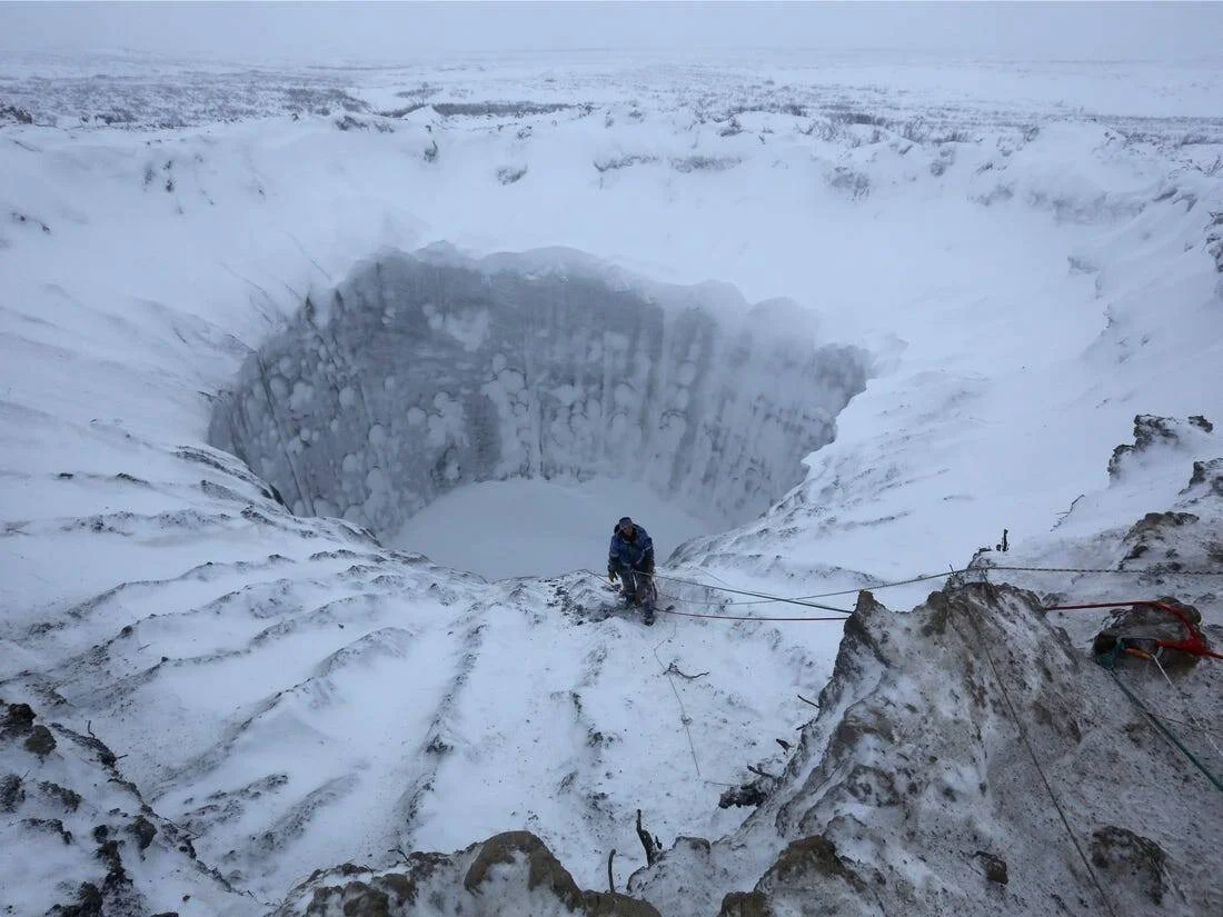 Un investigador sujeto con cuerdas se asoma al borde de un enorme cráter cilíndrico en la tundra ártica cubierta de nieve.