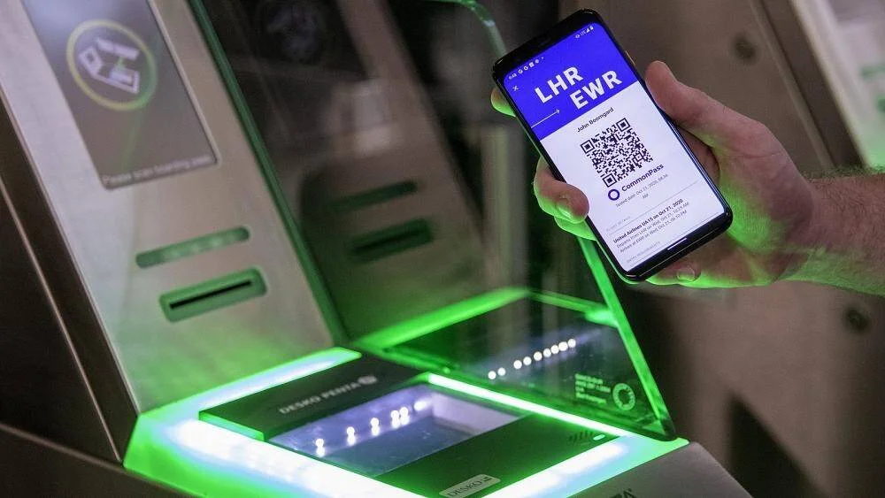 Mano sosteniendo un móvil con la app CommonPass mostrando un código QR para vuelo LHR-EWR ante un lector de documentos en aeropuerto.