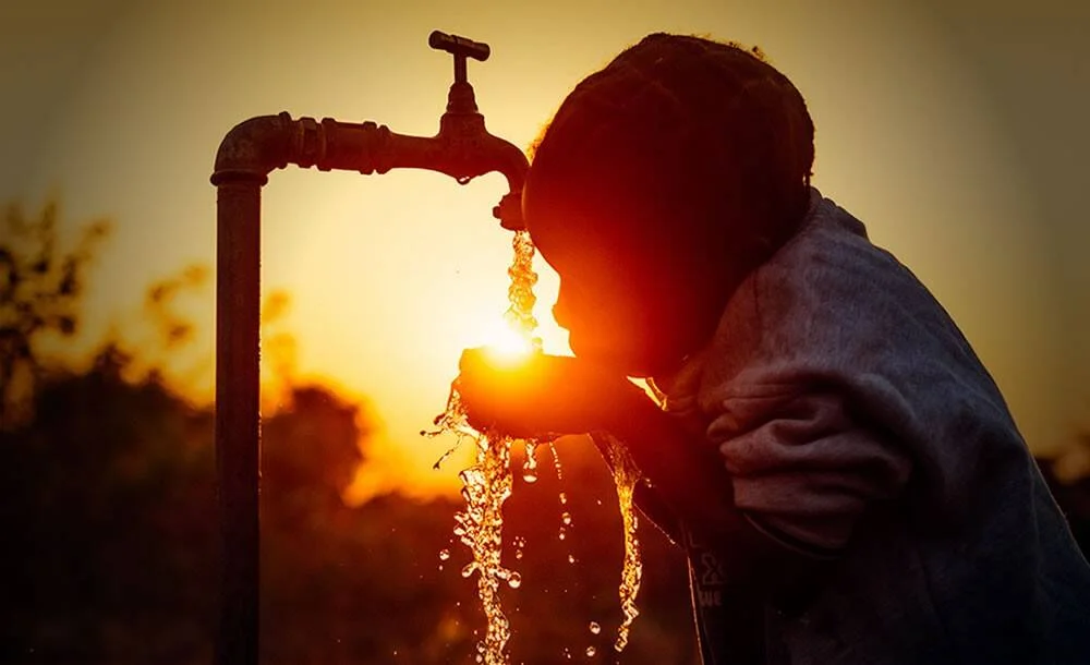 Niño en silueta bebe agua directamente de una canilla rural al aire libre, con el sol del atardecer de fondo.