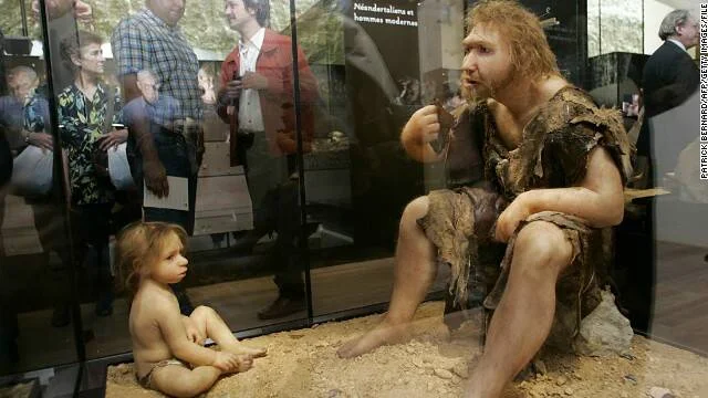 Figuras realistas de un neandertal adulto y un bebé expuestas en un museo, con visitantes reflejados en el vidrio al fondo.